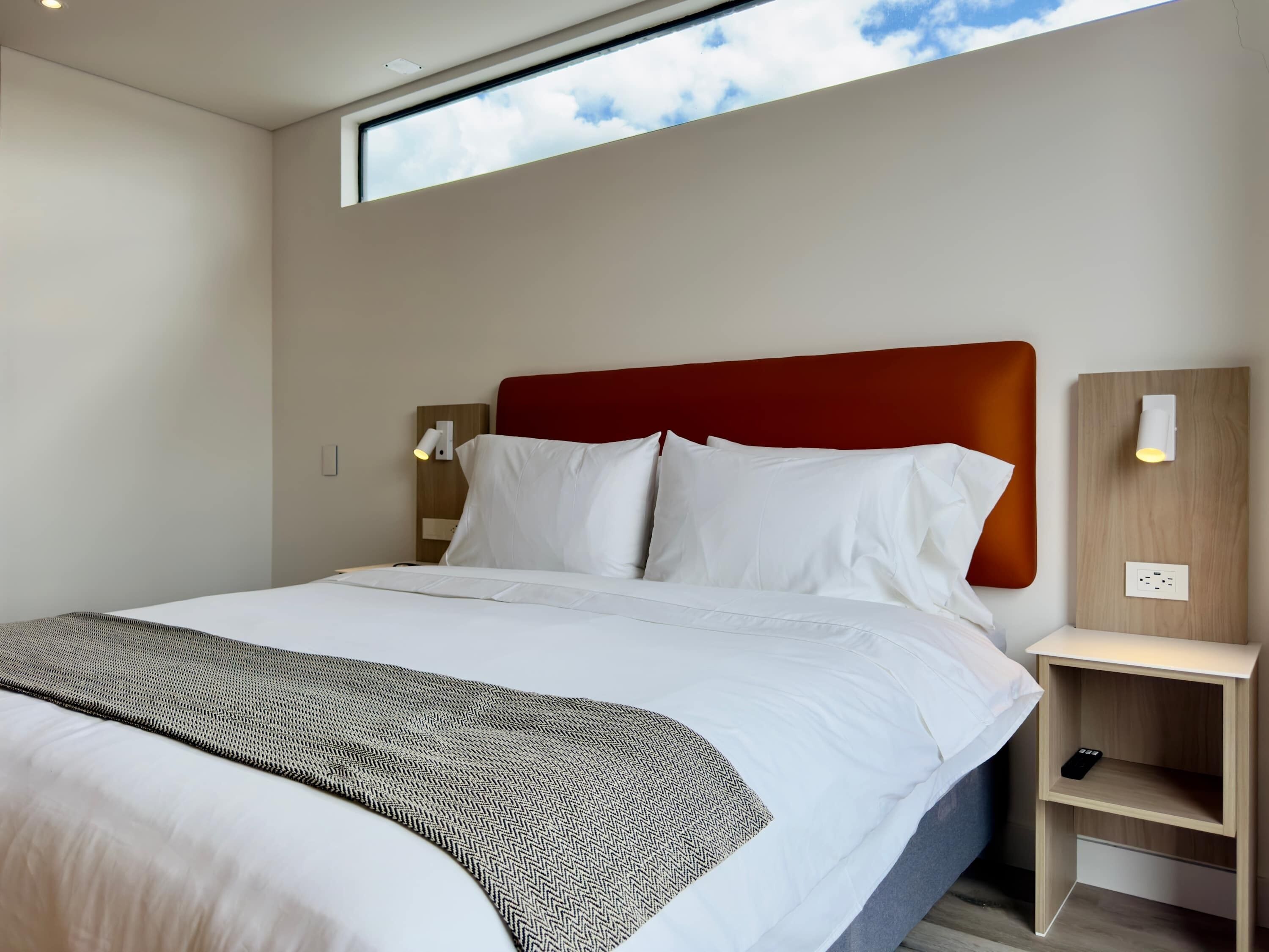 Une chambre d’hôtel moderne avec un grand lit, une tête de lit rouge, une table de chevet et une petite table d’appoint.