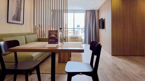 Une chambre d’hôtel avec un plancher en bois, un canapé vert, une table en bois avec deux chaises, un tableau encadré au mur, une bouteille d’eau et un téléviseur fixé au mur.