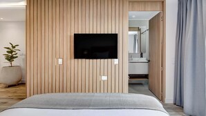Habitació d'hotel moderna amb un llit gran, una televisió de pantalla plana penjada a una paret de panells de fusta, una planta en test i un bany visible a través d'una porta oberta.