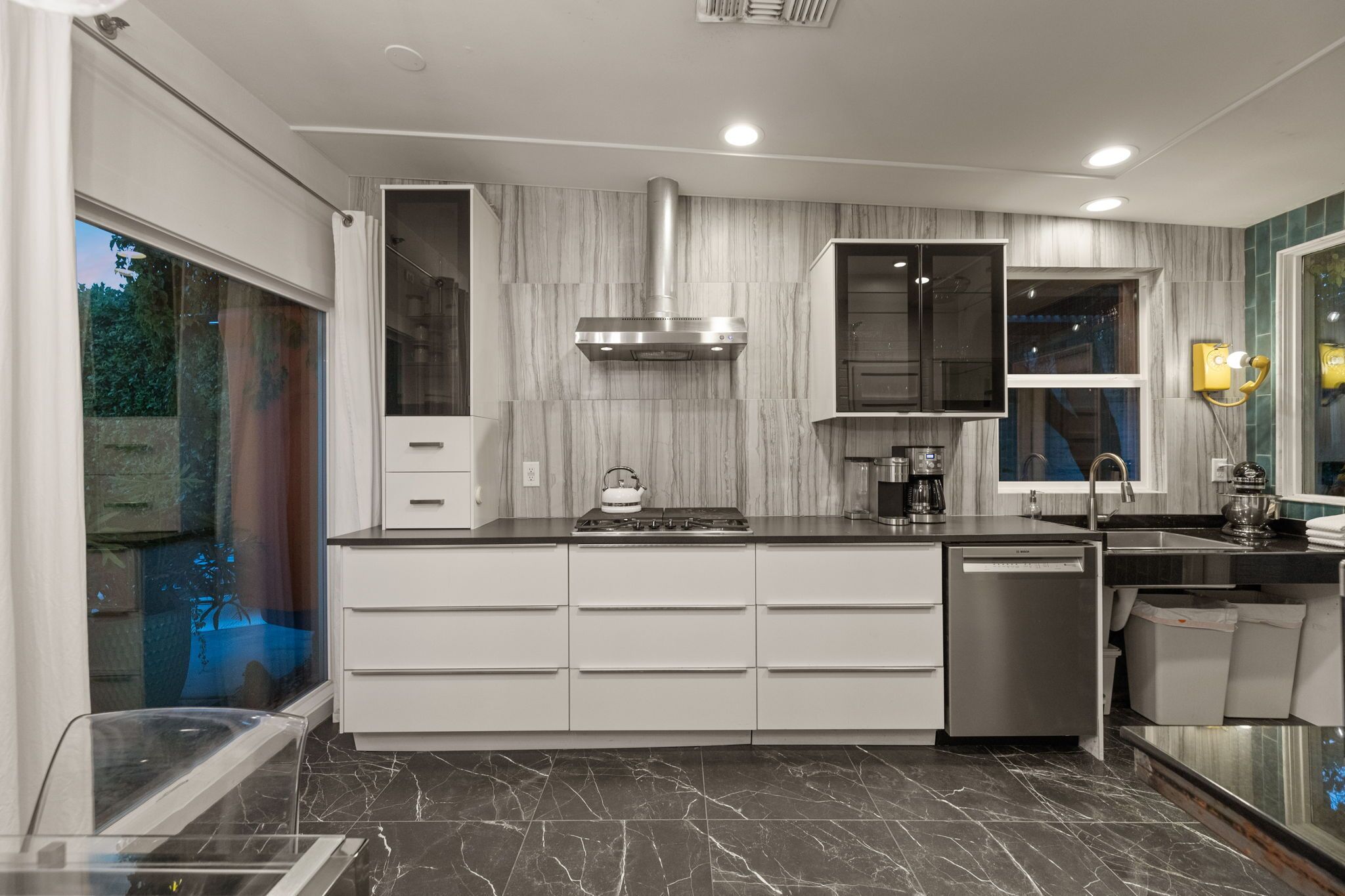 A modern kitchen with a built-in stove, a dishwasher, and a window with a view of greenery.