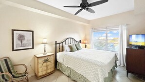 Une chambre à coucher comprenant un lit, un ventilateur de plafond, une table de chevet avec une lampe, une chaise, une commode, un téléviseur et une fenêtre donnant sur un beau paysage.