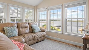 A living room with a beige leather sofa, patterned cushions, and large windows offering a view of a body of water and greenery. - Deep Water Access, Best Waterway Views, Pet-Friendly (Surf City)