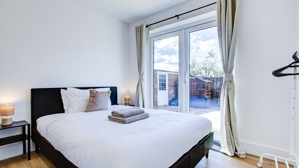A modern bedroom with a large bed, a nightstand with a lamp, and a view of the outside through a glass door. - Superb 4 Bed House with Driveway and Garden (Essex)