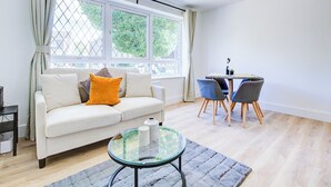 A living room with a sofa, a glass coffee table, a dining table with chairs, and a window with curtains. - Superb 4 Bed House with Driveway and Garden (Essex)