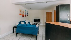 A modern living room with a blue sofa, a flat-screen TV, and a kitchen area with a black refrigerator. - Chic 3 Bed Flat on Harbour Way (Kent)