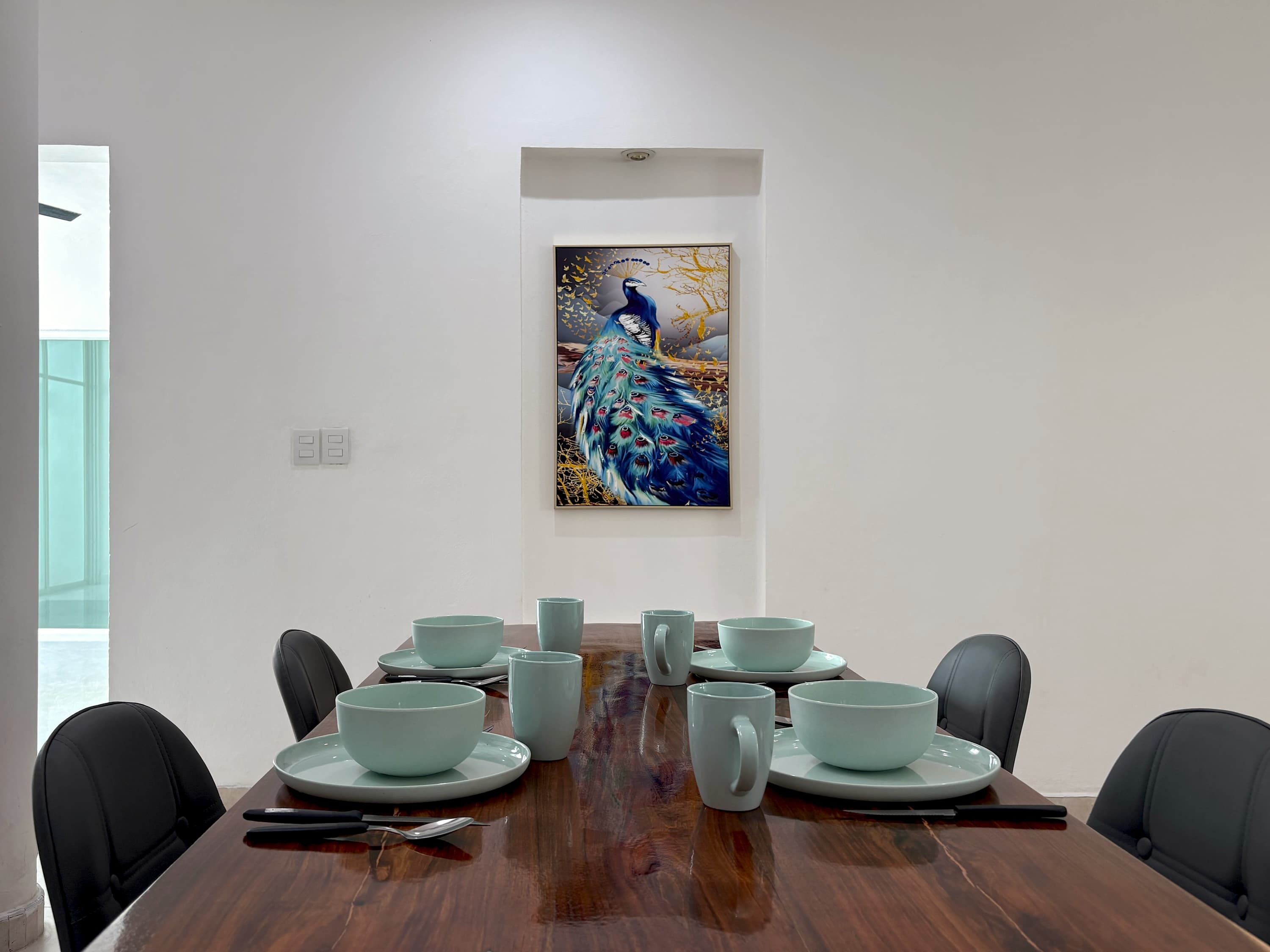 A dining room with a wooden table set for eight, surrounded by black chairs. A peacock painting is on the wall above the table.