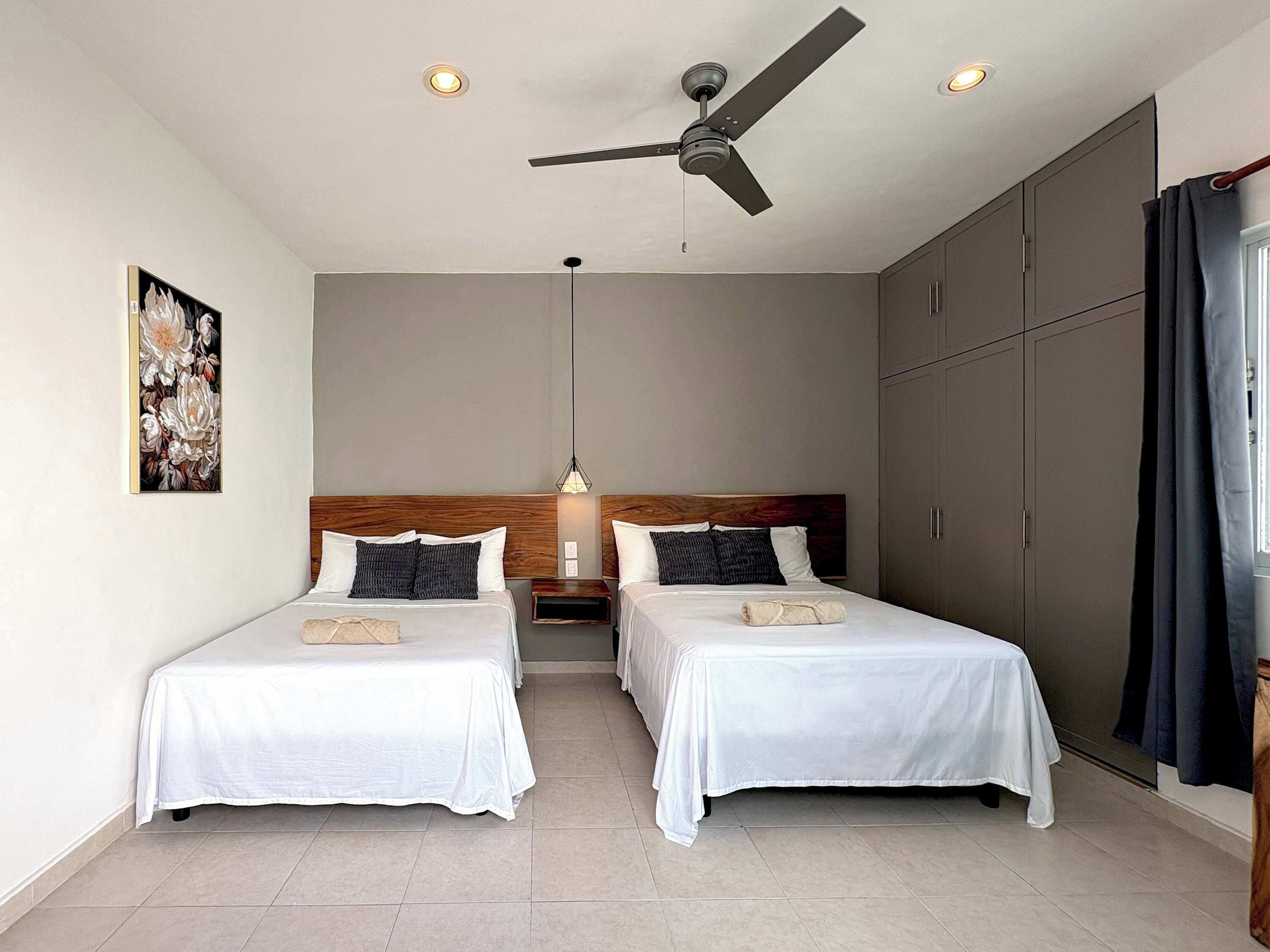 Une chambre d’hôtel avec deux lits simples, un ventilateur de plafond, un tableau au mur et une grande armoire.
