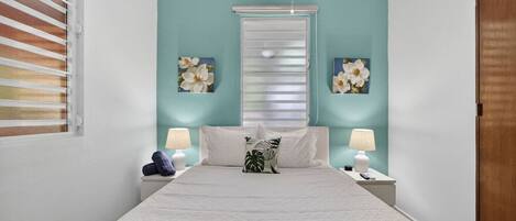 A bedroom with a bed, two nightstands, a window with blinds, and wall art.