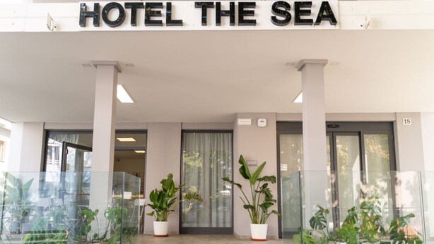 De ingang van Hotel The Sea met een modern ontwerp, met grote glazen deuren, plantenbakken en een helder zicht op het interieur.