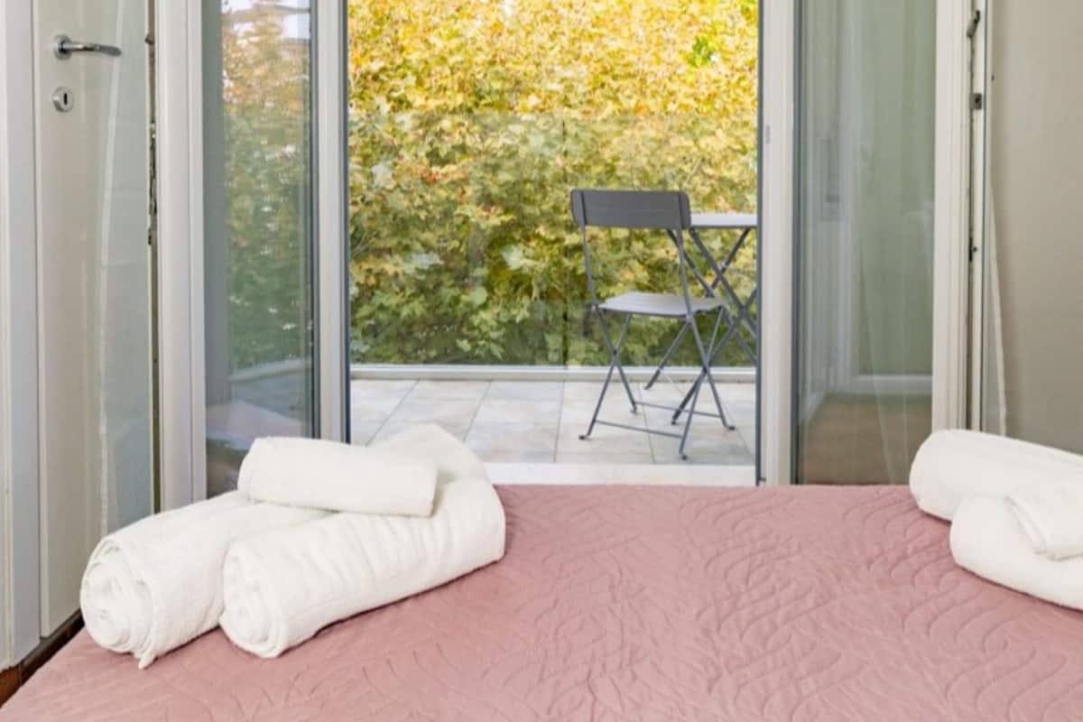 Een slaapkamer met een roze sprei, twee opgerolde handdoeken, een stoel en uitzicht op herfstbladeren door een glazen deur.