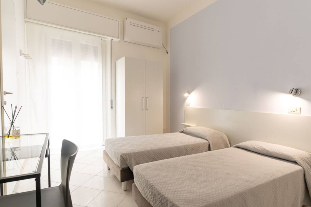 Hotelkamer met twee bedden, een bureau en een stoel. Er is een raam met gordijnen, een airconditioning en een wandlamp.