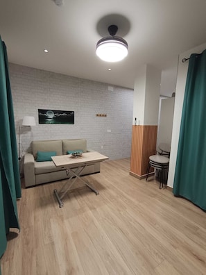 Ein modernes Zimmer mit einer weißen Ziegelwand, Holzboden, einer Couch mit türkisfarbenen Kissen, einem kleinen Tisch, einem Stuhl, einer Lampe und einer Deckenleuchte.
