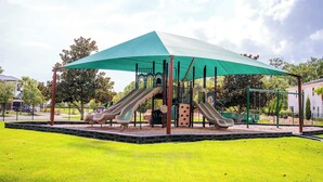 A playground with two slides and a climbing structure, covered by a green canopy, surrounded by a fenced grassy area. - First Floor Resort Condo Poolside Oasis (Kissimmee)