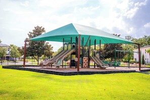 Ein Spielplatz mit zwei Rutschen und einer Kletterkonstruktion, überdacht von einem grünen Baldachin, umgeben von einem eingezäunten Rasenbereich.