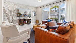 A modern living room with a white brick wall, a large flat-screen TV, a brown leather sofa, a wicker armchair, and a wooden coffee table. - Poolside Retreat at Reunion Resort Steps from the Water & Minutes to Disney (Kissimmee)