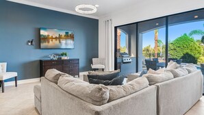 Ett modernt vardagsrum med en stor grå soffa, en platt-TV som är monterad på väggen och en matplats med utemöbler som syns genom stora glasdörrar.