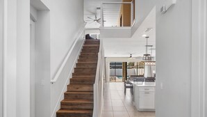 Una escalera conduce a un balcón en el segundo piso, con vista a una piscina, una zona de cocina con armarios blancos y un ventilador de techo.