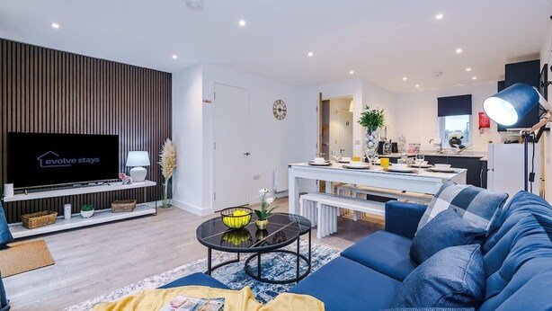 Uma sala moderna com televisão de tela plana, um sofá, uma mesa de centro e uma área de jantar com mesa e banco brancos.