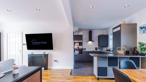 A modern living room with a flat-screen TV, a sofa, a dining table, and a kitchen area with a microwave and cabinets. - Game Room | Summer House | 3Bed Home In Stretford (Manchester)
