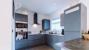 A modern kitchen with grey cabinets, a built-in oven, a range hood, and a window with blinds. - Game Room | Summer House | 3Bed Home In Stretford (Manchester)