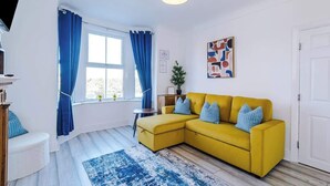 Una habitación con un sofá amarillo, un tapete azul y blanco, una puerta blanca y una ventana con cortinas azules.