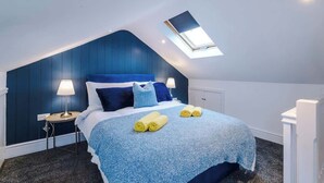 Ein Schlafzimmer mit schrägen Wänden, einem Oberlicht, einem Bett mit blauer Bettwäsche, zwei Nachttischlampen und einem kleinen Tisch mit einer Lampe.