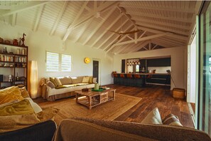 A spacious living room with a high ceiling, a cozy seating area, a kitchen with bar stools, and a large window providing natural light. - Villa Ravenala - Luxury Retreat with Breathtaking Caribbean Panoramic Views (Saint-Martin)