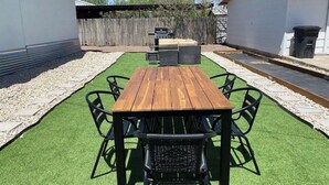 A wooden table with chairs in a backyard with artificial grass, a barbecue grill, and a shed in the background. - Charming 3-bedroom "The Rose House" in Marathon near Big Bend NP (Marathon)