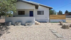 A modern single-story house with a flat roof, a blue door, and a small window. There are three metal containers on the ground, and a wooden fence is visible in the background. - Charming 3-bedroom "The Rose House" in Marathon near Big Bend NP (Marathon)