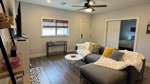 A modern living room with a grey sofa, a ceiling fan, and a window with blinds. - Charming 3-bedroom "The Rose House" in Marathon near Big Bend NP (Marathon)