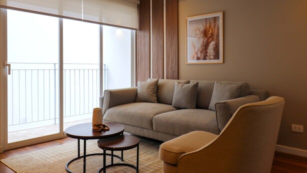 Una sala de estar moderna con un sofá gris, una mesa de centro redonda y un sillón beige. Hay un ventanal con persianas y una obra de arte enmarcada en la pared.