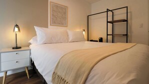 Una cama bien hecha con sábanas blancas, un plaid beige y una mesita de noche con una lámpara.