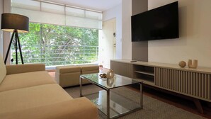 A modern living room with a sofa, a glass coffee table, a TV mounted on the wall, and a large window with a view of trees. - w* | Graceful 1BR with Balcony in Miraflores (Miraflores)