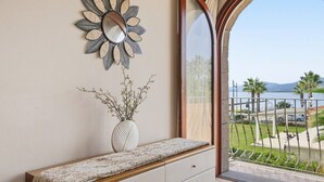 Una stanza con una panchina in legno, uno specchio decorativo, un vaso con rami e vista su un balcone con ringhiera e palme.