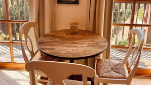 A round wooden table with a rustic finish, surrounded by four white chairs with woven cushions. A decorative wooden window frame with a gothic arch is centered above the table. - Ocean, Sunset, Mountain Views, Private Deck: Villa Floresco: Tuscan-like Getaway (Santa Barbara)