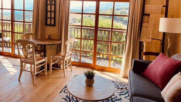 A room with a wooden floor, a round table with a plant, a sofa with cushions, and a view of greenery through large windows. - Ocean, Sunset, Mountain Views, Private Deck: Villa Floresco: Tuscan-like Getaway (Santa Barbara)
