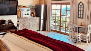 A bedroom with a large bed, a TV, a chandelier, a balcony with a table and chairs, and a kitchen area. - Ocean, Sunset, Mountain Views, Private Deck: Villa Floresco: Tuscan-like Getaway (Santa Barbara)
