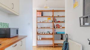 Een kleine keuken met een magnetron, houten kastjes en een boekenplank met diverse spullen.