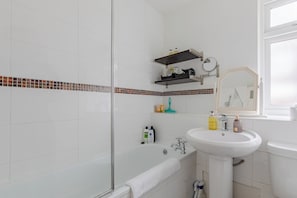 Ein Badezimmer mit einem weißen Waschbecken, einem Spiegel, einer Dusche und einer Toilette. Auf dem Waschbecken liegen Pflegeprodukte und am Boden ein Handtuch.