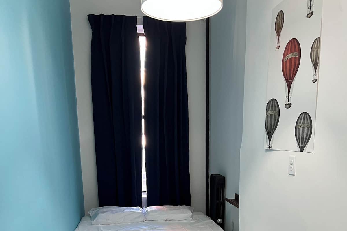A small, neatly made room with a bed, a chair, a lamp, and a wall with hot air balloon artwork.