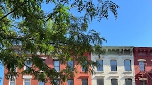 A row of red and white brick townhouses with fire escapes, parked cars, and a tree in the foreground. - East Harlem Hostel (New York)