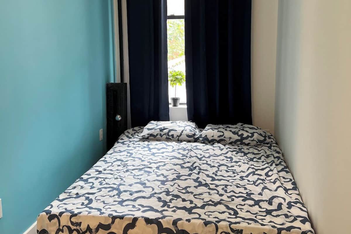 A small, single-bed room with a teal wall, a window with dark curtains, and a patterned bedspread.