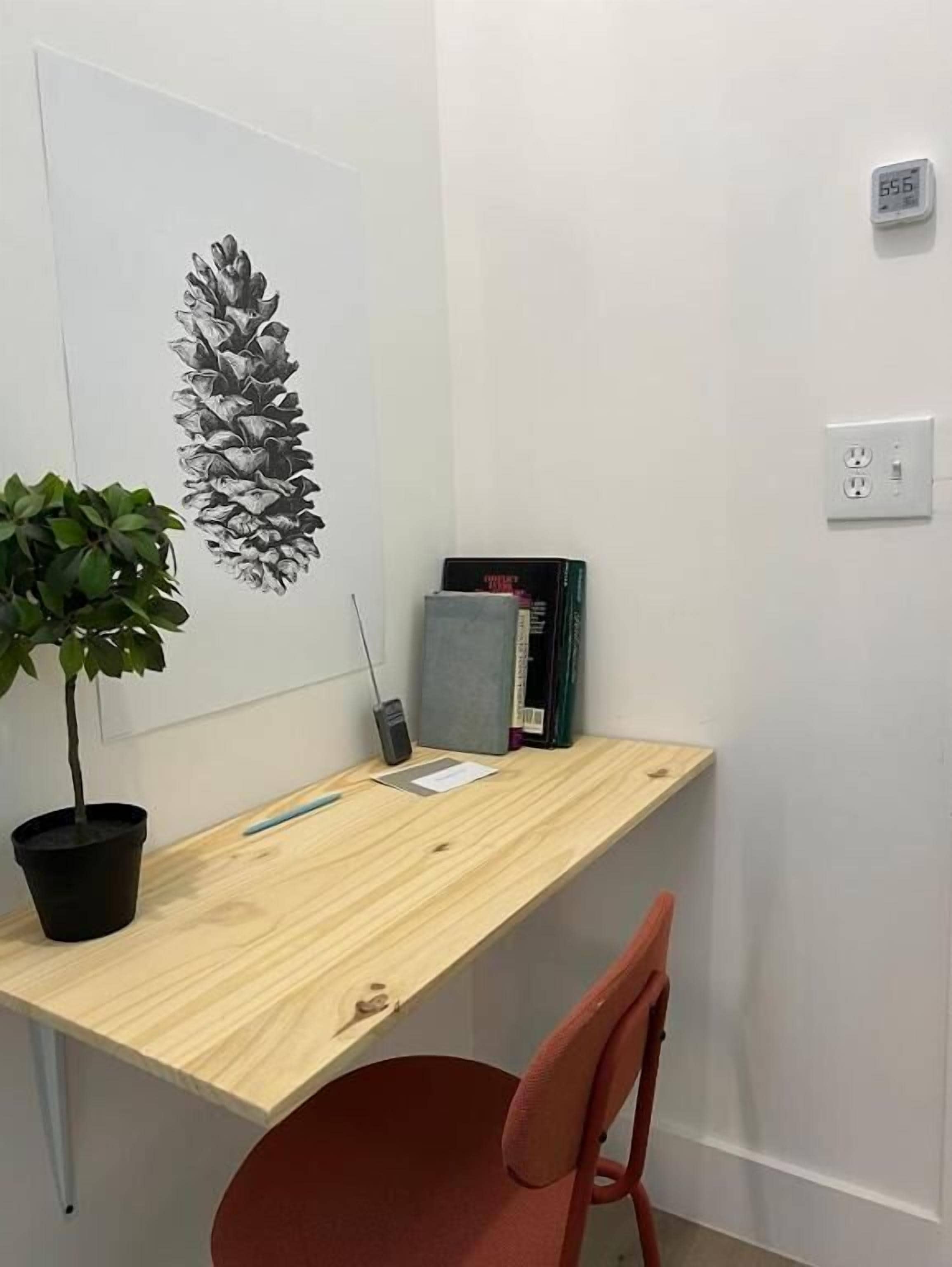A small, neatly organized office space with a wooden desk, a potted plant, a red chair, and a wall-mounted shelf with books and a phone.