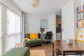 A modern living room with a green sofa, a wooden dining table, a small round table, a black office chair, and a kitchen visible in the background. - Bright and Trendy 1BD Canalside Flat in Hackney (Greater London)