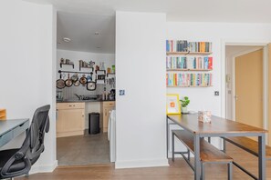 Une cuisine moderne avec un coin repas, comprenant une table et un banc en bois, une bibliothèque remplie de divers ouvrages et des ustensiles de cuisine suspendus au-dessus du plan de travail.