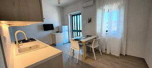 Una cocina compacta con un lavamanos, estufa y un refrigerador pequeño. Hay una mesa de comedor con sillas y un televisor montado en la pared.