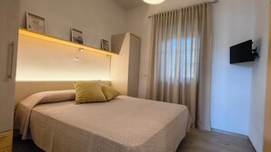 Un dormitorio con cama, armario, estante con fotografías, ventana con cortinas y televisor de pared.