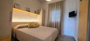 Une chambre à coucher comprenant un lit, une armoire, une étagère avec des photos, une fenêtre avec des rideaux et un téléviseur fixé au mur.
