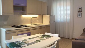 Cocina compacta con estufa a gas, pileta y armarios de madera. Hay una mesa de comedor con mantel decorado. Un sofá está ubicado cerca de una ventana con cortinas de gasa.