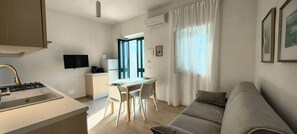 Une cuisine moderne équipée d’une cuisinière intégrée, un coin repas avec une table et des chaises, un canapé gris et un téléviseur fixé au mur.